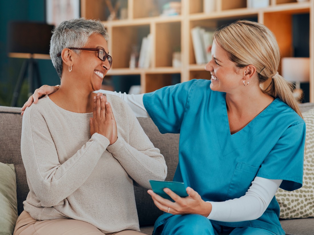 A female caregiver supports an older woman in her home while reviewing something on a small tablet.